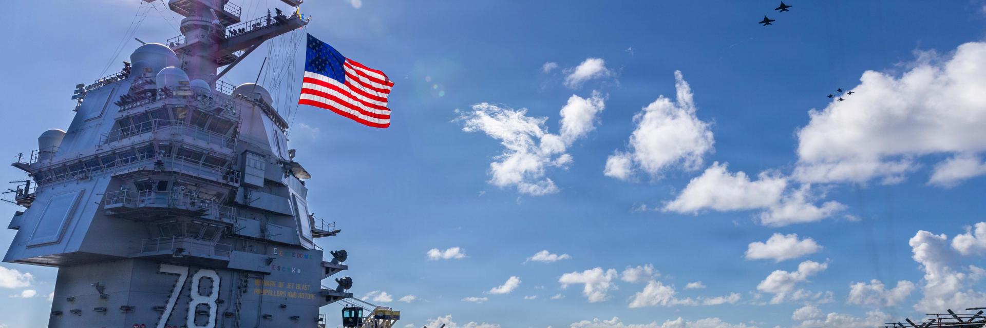 U.S. Navy aircraft carrier Gerald R. Ford under a bright sun, an American flag waving prominently. Several fighter jets are parked on the flight deck, while a formation of military aircraft flies overhead against a blue sky with scattered clouds. Credit photo:  U.S. Department of Defense. The appearance of U.S. Department of Defense (DoW) visual information does not imply or constitute DoW endorsement.