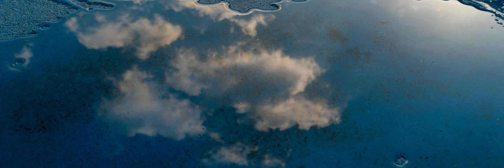 A cloudy sky is reflected in a shallow puddle on a dark blue concrete surface. The water's irregular edge and soft lighting create a calm, abstract mirror-like effect. Image credit: bxnjpg/Unsplash