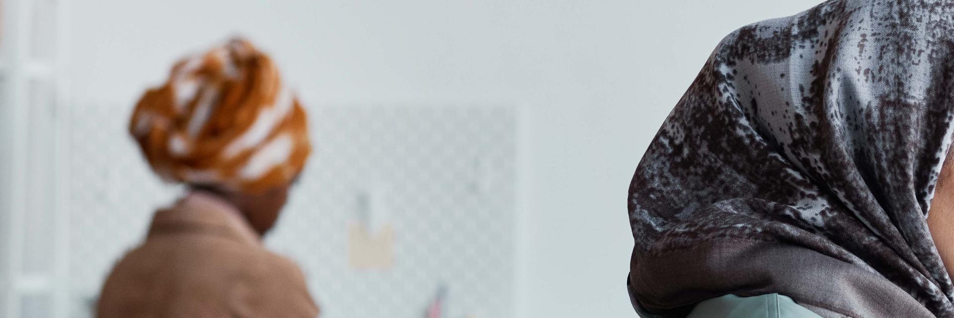 Two women are working in a minimal office setting, both wearing patterned headscarves. One is in the foreground facing right, partially out of frame, while the other sits in the background working on a laptop.