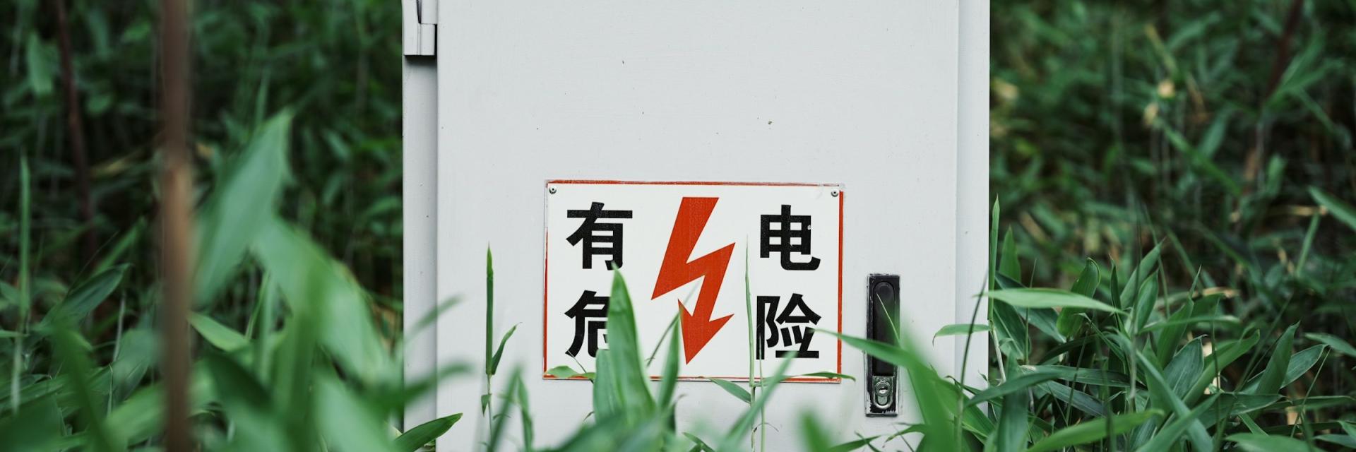 A white outdoor electrical utility box surrounded by tall green plants, with a warning sign featuring Chinese characters and a red lightning bolt indicating high voltage. Credit: Wenying Yuan via Unsplash