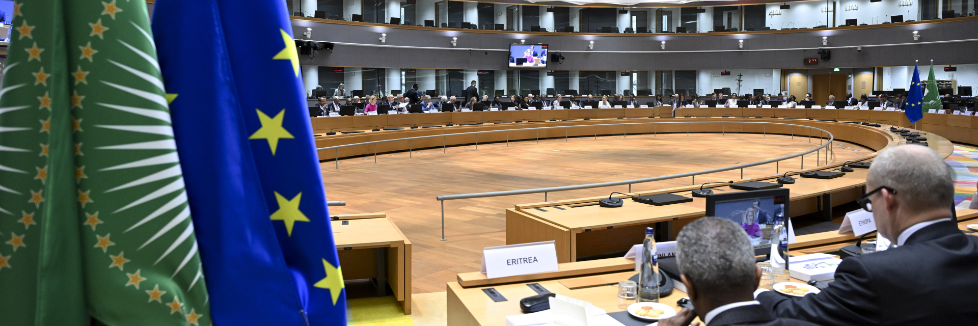 EU-African Union Ministerial meeting Roundtable. Two flags hang from the ceiling of a large room, where a group of people are seated at a long table. The people are engaged in a meeting, with some of them wearing ties. The room is filled with chairs, and a TV is mounted on the wall. (AI-generated text) | © European Union