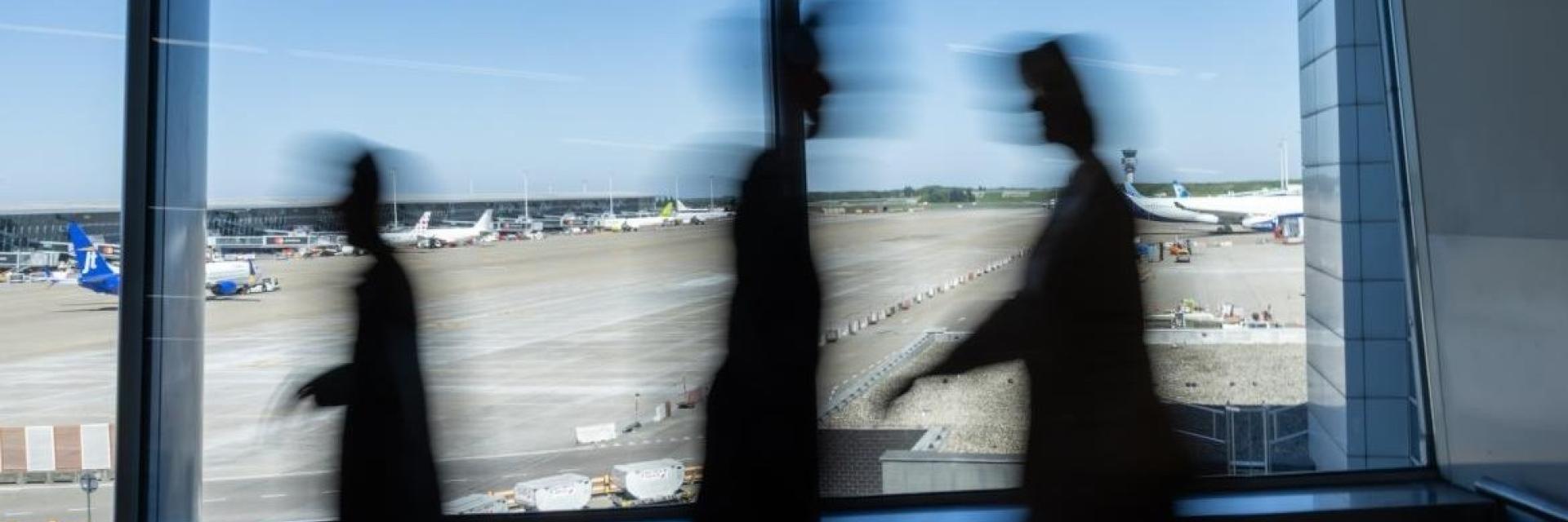 Three blurred people walking inside an airport terminal with airplanes visible outside through large windows. Image credit to the European Commission.