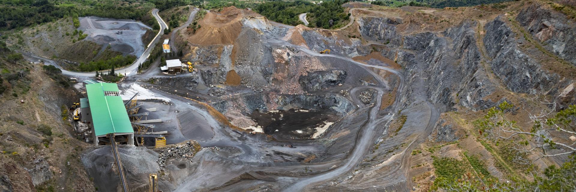 A large open-pit mining site with exposed earth, winding dirt roads and industrial structures including a green-roofed building surrounded by greenery and hills. Credit photo: Envato Elements