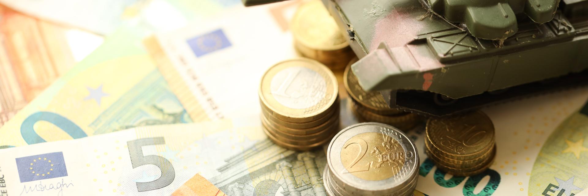 A toy military tank sits atop a pile of Euro notes and coins.
