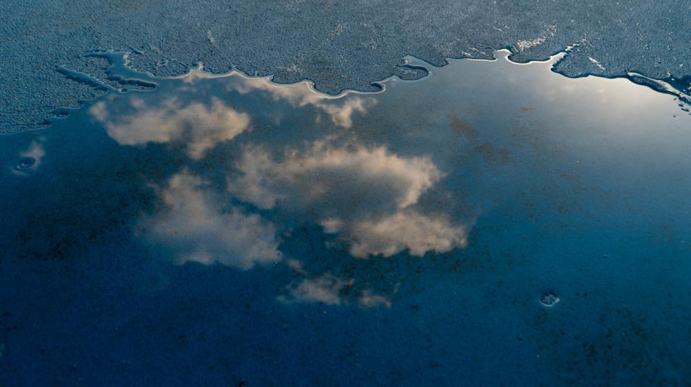 A cloudy sky is reflected in a shallow puddle on a dark blue concrete surface. The water's irregular edge and soft lighting create a calm, abstract mirror-like effect. Image credit: bxnjpg/Unsplash
