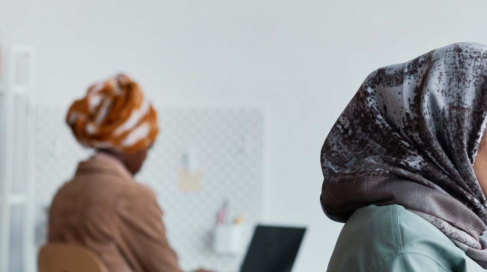 Two women are working in a minimal office setting, both wearing patterned headscarves. One is in the foreground facing right, partially out of frame, while the other sits in the background working on a laptop.