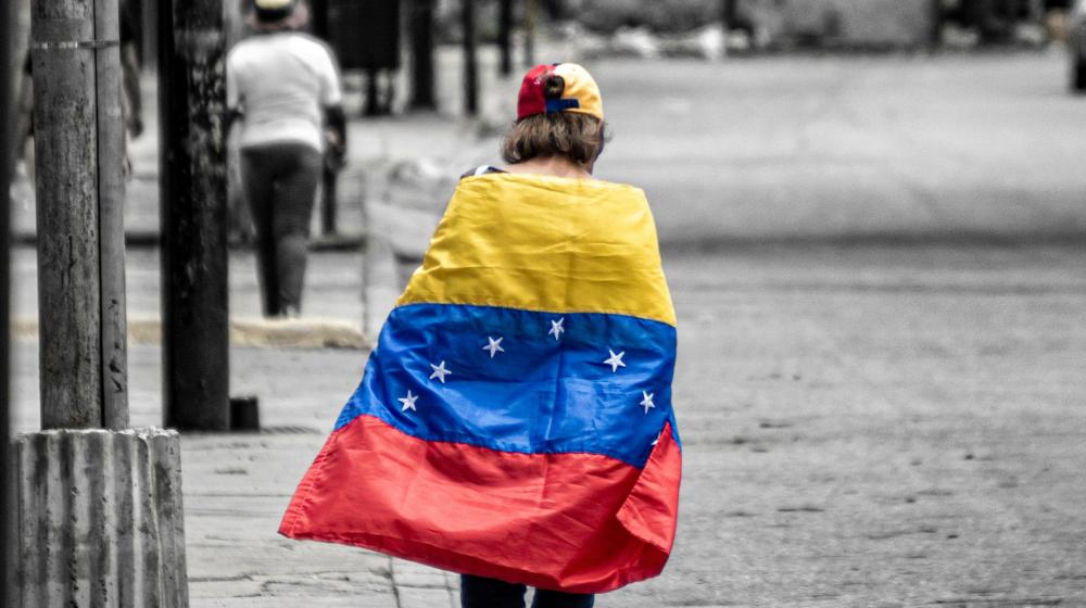 Boy walking in the street with a Venezuelan flag draped over his shoulders. Photo by Andrés Silva on Unsplash 