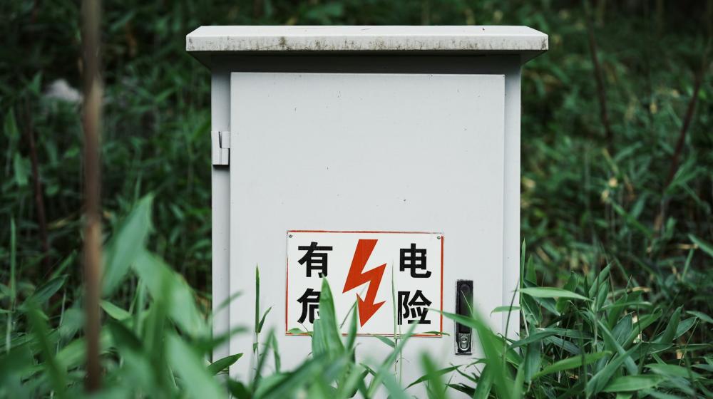 A white outdoor electrical utility box surrounded by tall green plants, with a warning sign featuring Chinese characters and a red lightning bolt indicating high voltage. Credit: Wenying Yuan via Unsplash