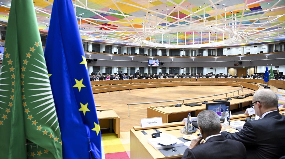 EU and African Union Ministerial meeting Roundtable. Two flags hang from the ceiling of a large room where a group of people are seated at a long table. The people are engaged in a meeting. The room is filled with chairs and a TV is mounted on the wall. Credit photo: European Union