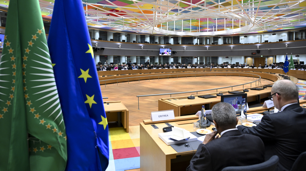 EU-African Union Ministerial meeting Roundtable. Two flags hang from the ceiling of a large room, where a group of people are seated at a long table. The people are engaged in a meeting, with some of them wearing ties. The room is filled with chairs, and a TV is mounted on the wall. (AI-generated text) | © European Union