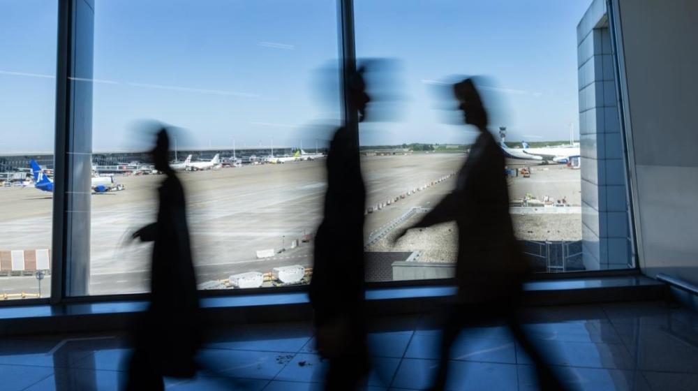Three blurred people walking inside an airport terminal with airplanes visible outside through large windows. Image credit to the European Commission.