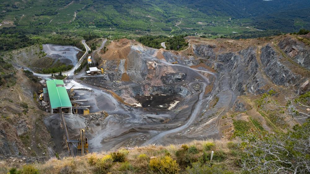 A large open-pit mining site with exposed earth, winding dirt roads and industrial structures including a green-roofed building surrounded by greenery and hills. Credit photo: Envato Elements