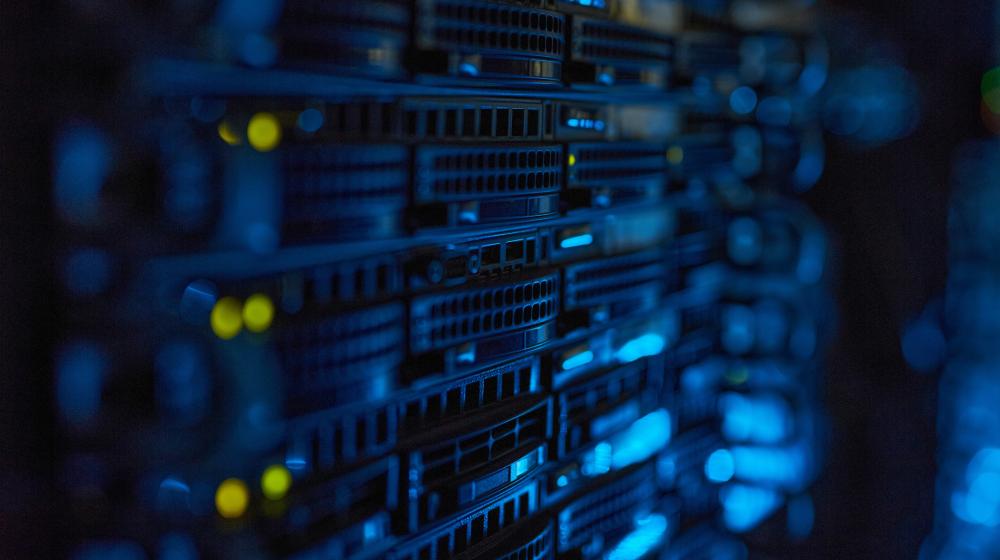 Rows of server racks with blue lighting and small yellow indicator lights in a dark data center environment. Image credit to Envato Elements.