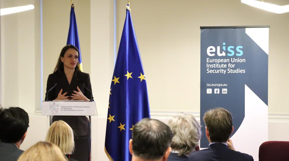 Romania Foreign Minister Oana Toiu standing on stage in front of EU and Romanian flags and EUISS banner. Credit photo: EUISS