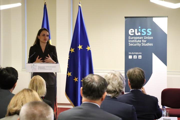 Romania Foreign Minister Oana Toiu standing on stage in front of EU and Romanian flags and EUISS banner. Credit photo: EUISS